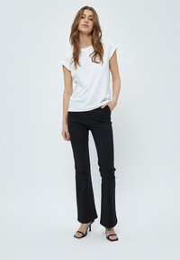 T-shirt bianco a maniche corte abbinato a pantaloni neri a campana e sandali neri con tacco, su uno sfondo grigio chiaro.