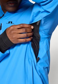 Casaco azul com um bolso lateral com fecho e interior em malha. O design inclui um colarinho alto e detalhes em tecido preto.