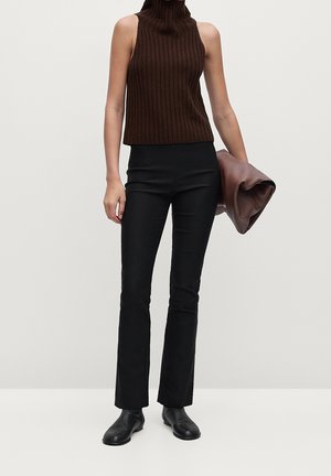 Vrouw met een mouwloze bruine coltrui, zwarte strakke broek en zwarte schoenen, die een gevouwen bruine leren clutch vasthoudt.