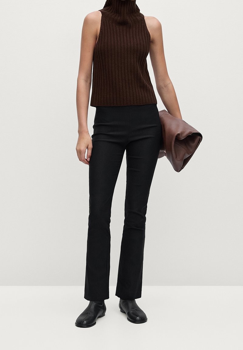 Femme portant un haut marron sans manches à col roulé, un pantalon noir ajusté et des chaussures noires, tenant une pochette en cuir marron pliée.