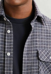 Camată din flanel gri în carouri, cu nasturi albi, un buzunar pe piept și un guler marin sub. Prim-plan al texturii și modelului țesăturii.