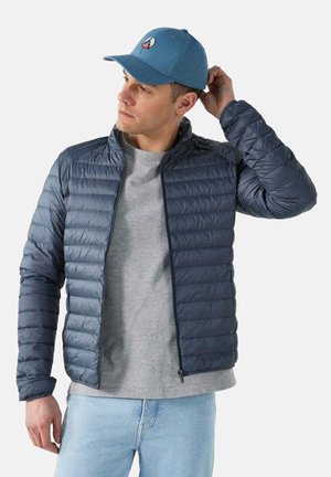 Marineblauwe pufferjack met een gestikt ontwerp, staande kraag, ritssluiting aan de voorkant, gecombineerd met een grijze t-shirt en lichtblauwe jeans, gedragen met een blauwe pet.