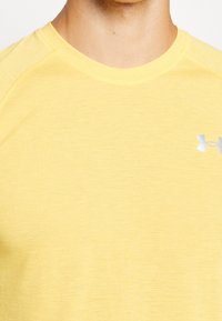 Žluté sportovní tričko s kulatým výstřihem, vyrobené z lehkého materiálu, s malým stříbrným logem na hrudi.