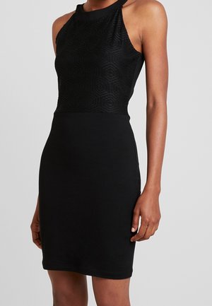 Femme portant une robe noire sans manches avec un motif géométrique texturé sur le buste supérieur, une taille cintrée et une jupe unie sur fond gris.