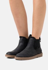 Svarta ankelstövlar i mocka med en mjuk krage kantad med päls och elastiska sidopaneler; de har en texturerad gummisula.