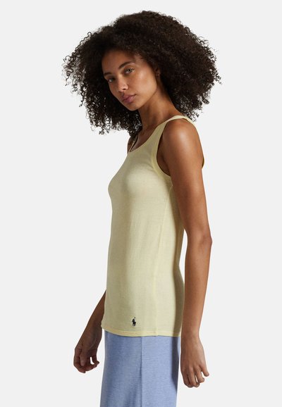 Femme aux cheveux bouclés portant un haut sans manches jaune pâle et un pantalon bleu clair, debout de profil sur un fond uni.