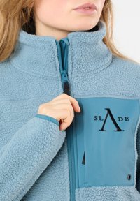 Giacca in pile di colore azzurro chiaro con superficie testurizzata, chiusura frontale con zip e tasca sul petto con logo nero e dettagli a contrasto.