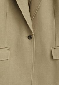 Blazer beige sur mesure avec revers crantés, bouton noir central et deux poches avant à rabat.