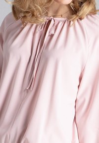 Jasnoróżowa bluzka z marszczonym okrągłym dekoltem i ozdobnym wiązaniem. Wykonana z gładkiej tkaniny, z długimi, zwiewnymi rękawami.