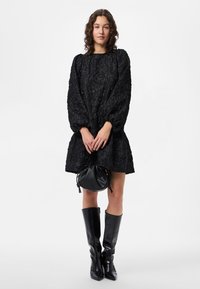 Crna haljina s teksturom, bufaste rukave i raširenim rubom; uparena s crnim čizmama do koljena i malom okruglom crnom torbom.