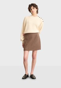 Suéter de rayas crema con botones en los hombros, combinado con una mini falda de espiga marrón. La modelo lleva zapatos de charol negros. Fondo neutro.