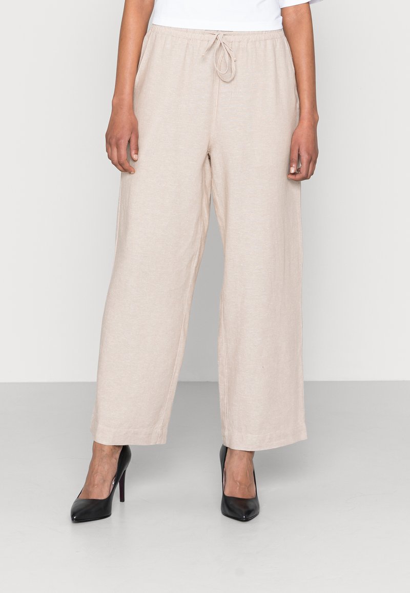 Beige vida byxor med elastisk midjeband och dragsko, tillverkade av mjukt, lättviktigt tyg. Kombineras med svarta klackskor.