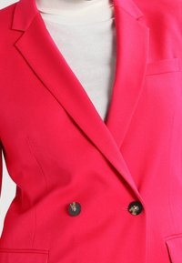 Fuchsiové sako se strukturálním designem, dvojitým zapínáním na knoflíky a hladkou látkou. Má dvě kapsy a výrazné klopy.