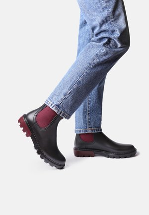 Black ankle boots with maroon elastic side panels and textured rubber soles, paired with faded blue jeans. Smooth leather finish.