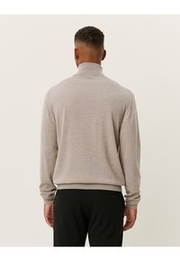 Beige Strickpullover mit hohem Kragen, langen Ärmeln und taillierter Silhouette, der eine subtile Textur und eine glatte Oberfläche auf der Rückseite aufweist.