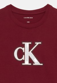 T-shirt Calvin Klein Jeans di colore marsala con logo CK bianco scintillante centrato sul petto, taglia 8 (128) all'interno del colletto.