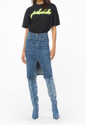Kvinde iført sort t-shirt med neontekst, blå denim nederdel med frontslids og matchende høje denimstøvler.