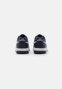 Námořnické modré sportovní boty s bílými akcenty, hladký syntetický materiál, polstrovaný límec a texturovaná gumová podrážka. Logo "New Balance" na patě.