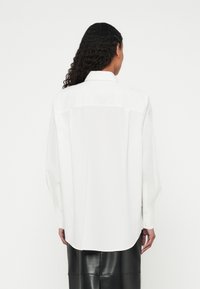 Blusa bianca dal taglio morbido, maniche lunghe e colletto appuntito. Presenta una spalla posteriore e polsini con bottoni. Finitura in tessuto liscio.