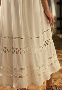 Vit maxiklänning med delikata spets- och broderidetaljer, med en flödande siluett och texturerad tyg, som framhäver intrikata mönster.