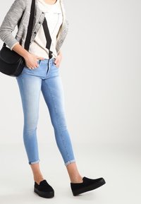 Svetle modré úzké džíny s roztrhaným lemem, kombinované se šedým cardiganem a grafickým tričkem, doplněné černými nazouvacími botami.