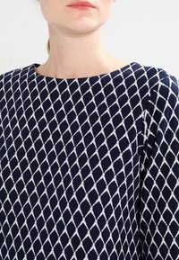 Marineblaues Strickoberteil mit einem weißen Rautenmuster, weitem Halsausschnitt und langen Ärmeln. Glatte Textur, lässiges Design für vielseitige Tragemöglichkeiten.