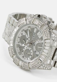 Montre-bracelet argentée avec un cadran texturé scintillant, trois sous-cadrans et un large bracelet orné de strass rectangulaires et ronds.