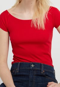 Röd kortärmad topp med vid halsringning, tillverkad av ett slätt tyg, matchad med mörkblå högmidjade jeans med knappring och dragkedja.