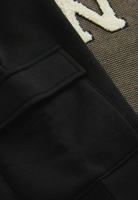 Mustapintainen kangas, jossa on teksturoitu pinta ja läpällä varustettu tasku, yhdistettynä beige-kermanväriseen houndstooth-kuvioon, jossa on kohotetut kirjaimet.