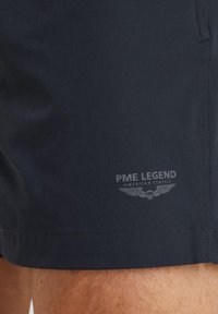 Shorts de bain bleu marine foncé en tissu léger. Présentent des coutures cousues et un logo avec des ailes dans une teinte plus claire à l'avant.