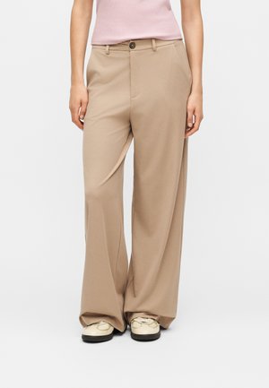 Femme portant un pantalon beige à jambes larges, un haut sans manches rose clair et des baskets crème, debout devant un fond blanc.