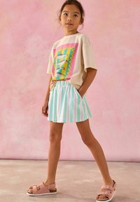 T-shirt beige clair avec un graphique de poisson coloré, associé à une jupe rayée menthe et blanche, et des sandales roses avec des straps ajustables.