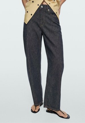 Person trägt dunkelblaue Straight-Leg-Jeans mit einem beigen, geknöpften Top und schwarzen flachen Sandalen mit Riemchen, steht vor einfachem Hintergrund.