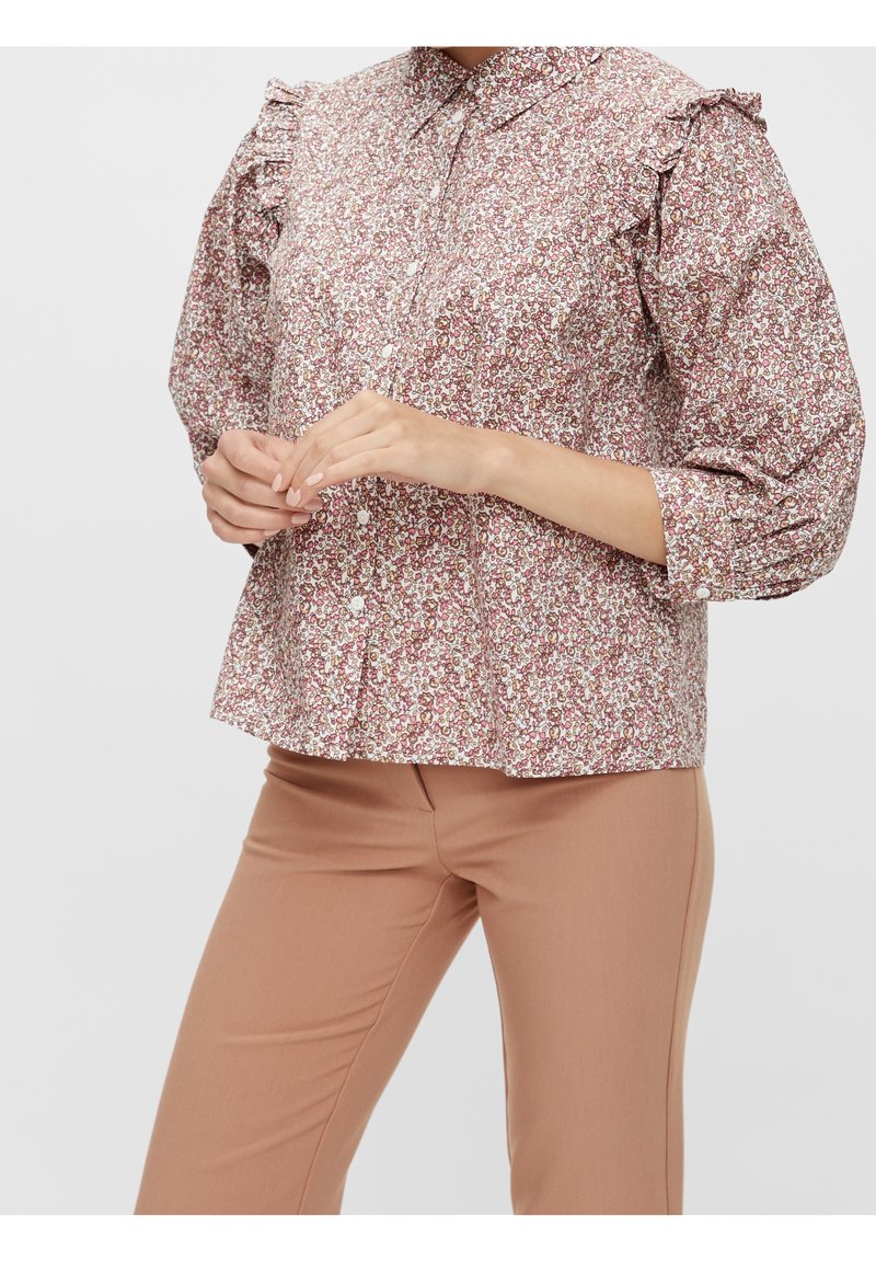 Blusa floreale con spalle arricciate, caratterizzata da un motivo rosa e crema, apertura con bottoni e colletto rotondo. Abbinata a pantaloni beige.
