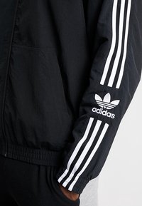 Giacca a vento nera con design a tre strisce bianche Adidas sulle maniche e logo sul polsino; vita elastica e texture liscia.
