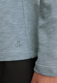 Gaiši zilā garām piedurknēm krekls ar teksturētu audumu, kam ir neliela 'M OP' logo apakšmalā un piedurkņu dizains ar aprocēm.