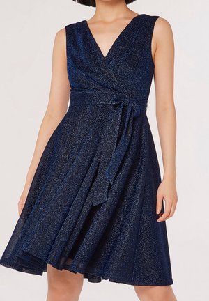 Ærmeløs navyblå glitrende slå-om-kjole med V-hals og bindebælte i taljen, båret af en kvinde med kort bob-frisure.