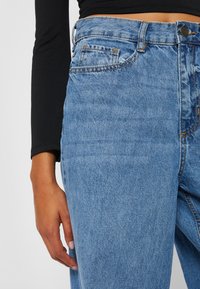 Light blue denim jeans with a high waist, featuring five pockets, orange stitching, and a smooth texture. Hand resting on the leg.