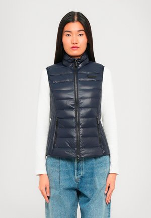 Vrouw in een marineblauwe gewatteerde mouwloze ritsvest over een wit longsleeve shirt en losse blauwe jeans, staand tegen een effen achtergrond.