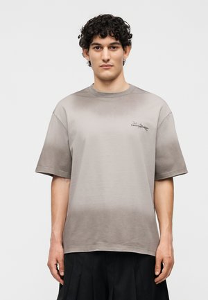 Νέος άντρας με σγουρά μαλλιά, φοράει άνετο γκρι μπλουζάκι με διαβάθμιση και μαύρο παντελόνι σε απλό ανοιχτό φόντο.