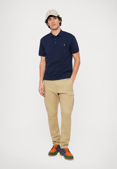 Mornarska polo majica, bež cargo hlače in večbarvni čevlji. Kapa je svetlo siva. Ima teksturirano tkanino in vidne šive.