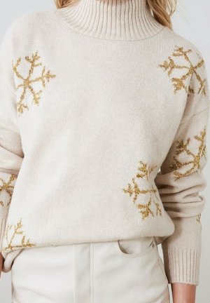 Cremefarbener Rollkragenpullover mit goldglitzernden Schneeflockenmustern, getragen zu elfenbeinfarbenen Hosen.
