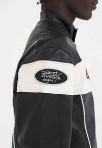 Giacca di pelle nera con un pannello bianco sulla spalla e un logo circolare. Il design include un colletto alto e dettagli di cucitura a contrasto.