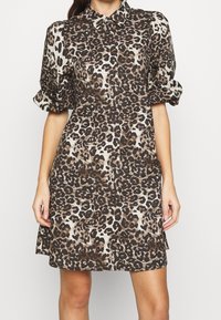 Kvinna som bär en klänning med korta ärmar och knappar framtill, med ett mönster i brunt och svart leopardtryck, krage och lätt puffade ärmar.