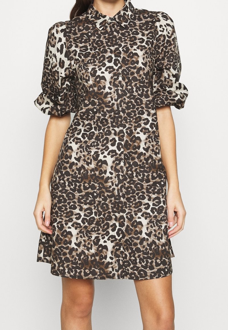 Kvinna som bär en klänning med korta ärmar och knappar framtill, med ett mönster i brunt och svart leopardtryck, krage och lätt puffade ärmar.