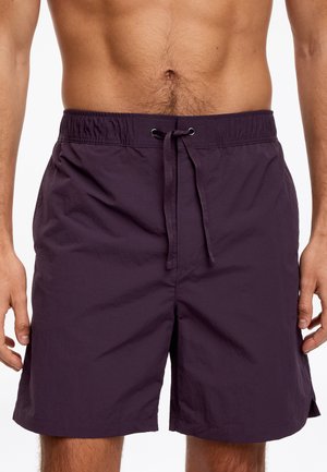 Homme portant un short de bain violet foncé arrivant à mi-cuisse avec une taille élastique et un cordon, debout avec les mains détendues sur les côtés.