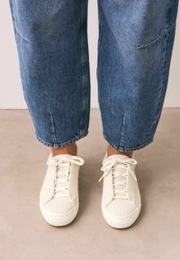 Des baskets blanches avec un dessus en tricot texturé, un bout rond et une semelle en caoutchouc plate, associées à un jean en denim bleu à coupe décontractée.