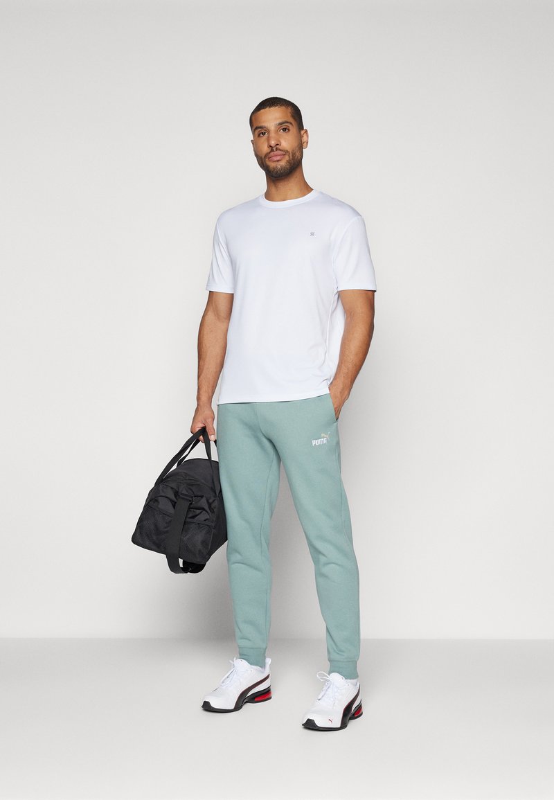 T-shirt bianco a maniche corte, pantaloni della tuta verde chiaro e borsa da viaggio nera. Le scarpe sono bianche con dettagli rossi e neri. Loghi semplici visibili.