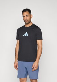 Melna īsām piedurknēm T-kreklu ar gaiši zilu Adidas logotipu. Audums šķiet gluds, apvienots ar gaiši zilu šortiem. Vienkāršs dizains un piegriezums.