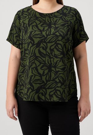 Vrouw draagt een groene top met korte mouwen met zwarte abstracte bladpatronen en een zwarte broek, staand tegen een effen achtergrond.